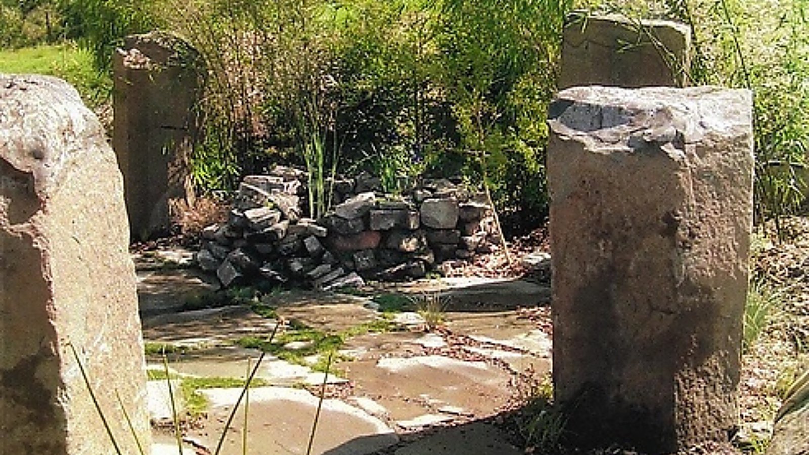 Weathered stone pillars beside a broken stone well feature, softened by naturalistic planting